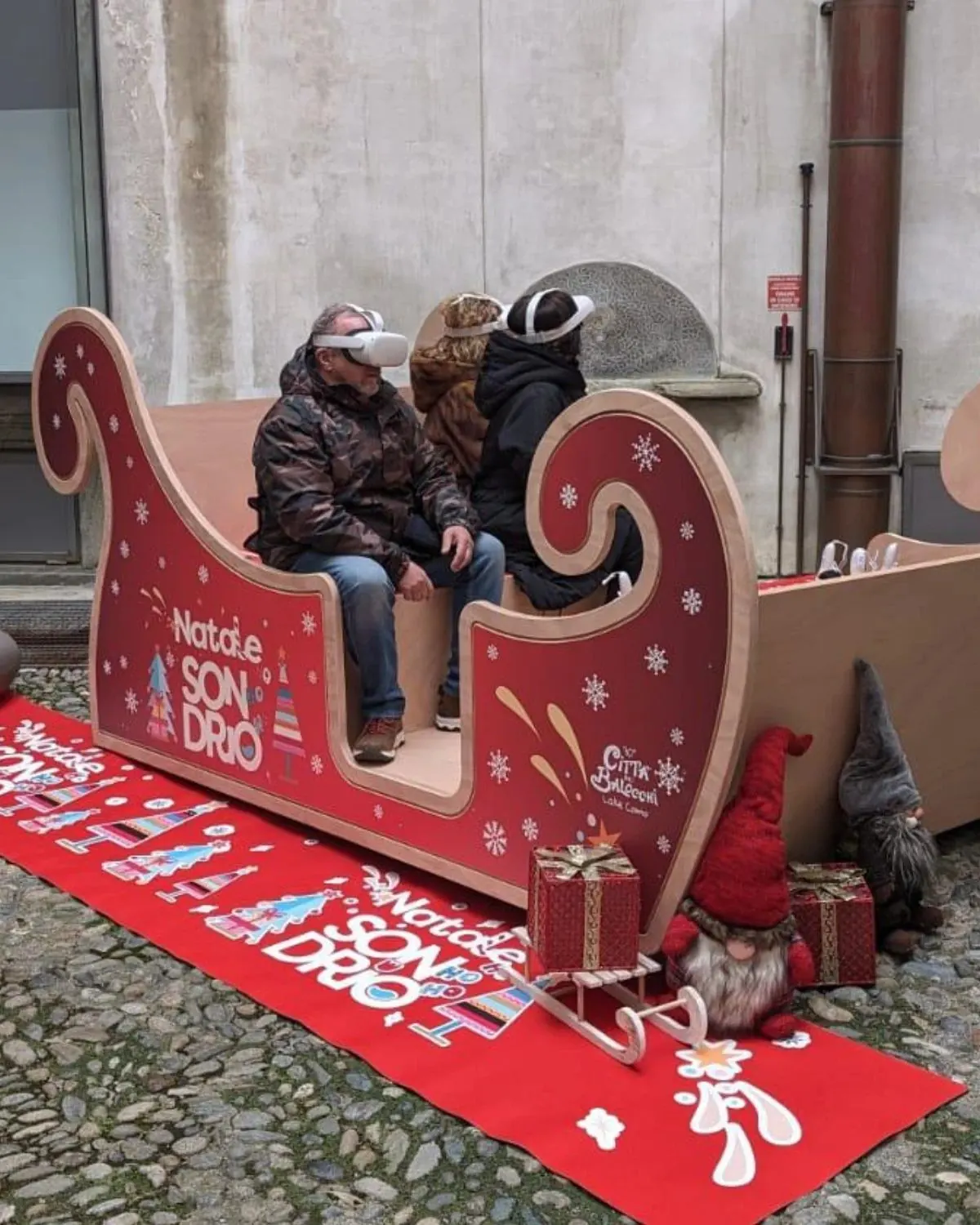 Tre persone con il visore sedute su una slitta di Babbo Natale, in un cortile allestito per le feste